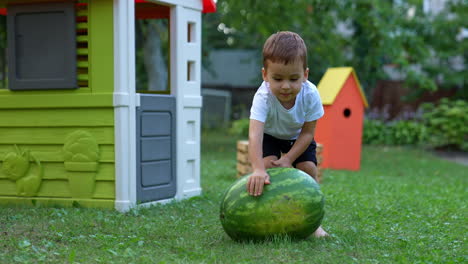 persistent caucasian baby boy pushing a big water-melon by the grass. happy child playing at the backyard.