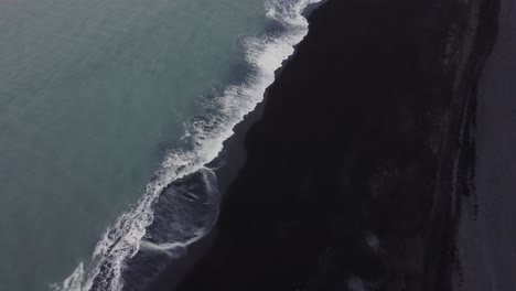 Aufsteigende-Luftdrohnenaufnahme-Wunderschöner-Blauer-Wellen,-Die-An-Einem-Schwarzen-Sandstrand-In-Island-Zusammenbrechen,-Und-Riesiger-Felsformationen-In-Der-Ferne