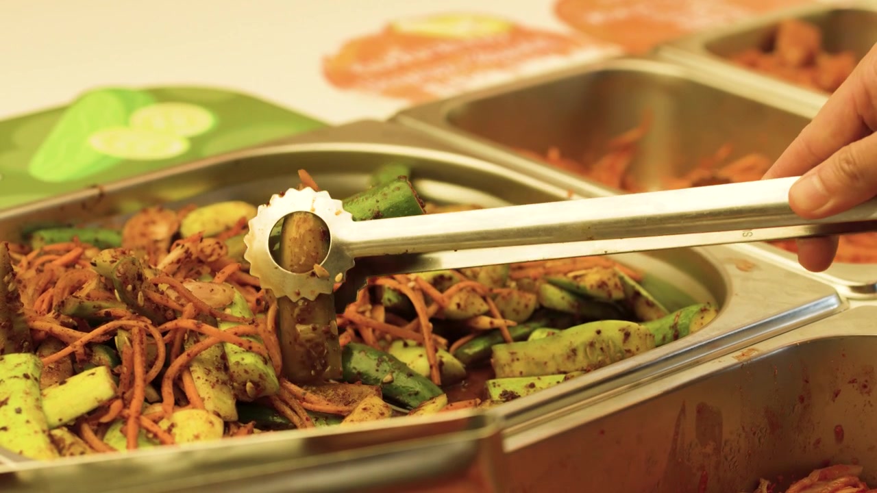 Pinças de metal pegam kimchi de pepino fermentado de uma bandeja de buffet de aço inoxidável em um restaurante