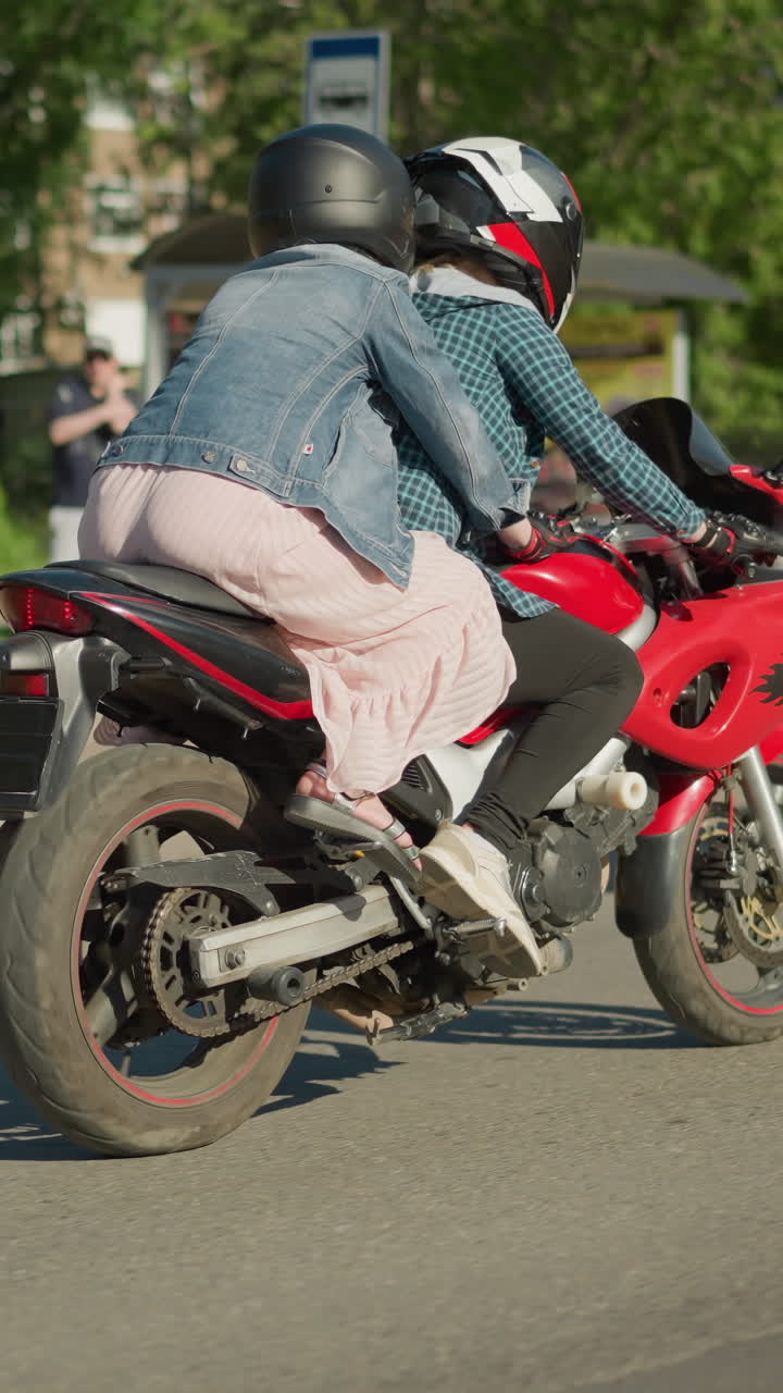 두 여인이 도시 도로를 가로질러 전기 자전거를 천천히 타고, 승객이 라이더를 붙잡고, 사람들의 흐릿한 시야, 주차된 자동차, 그리고 높은 나무가 배경에서 보인다.