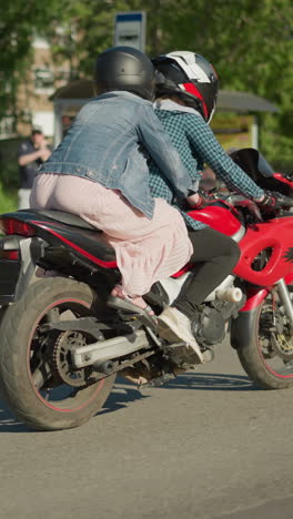 dos mujeres viajan lentamente en una bicicleta eléctrica a través de una carretera urbana, el pasajero se aferra al jinete, se ven vistas borrosas de personas, coches estacionados y árboles altos en el fondo