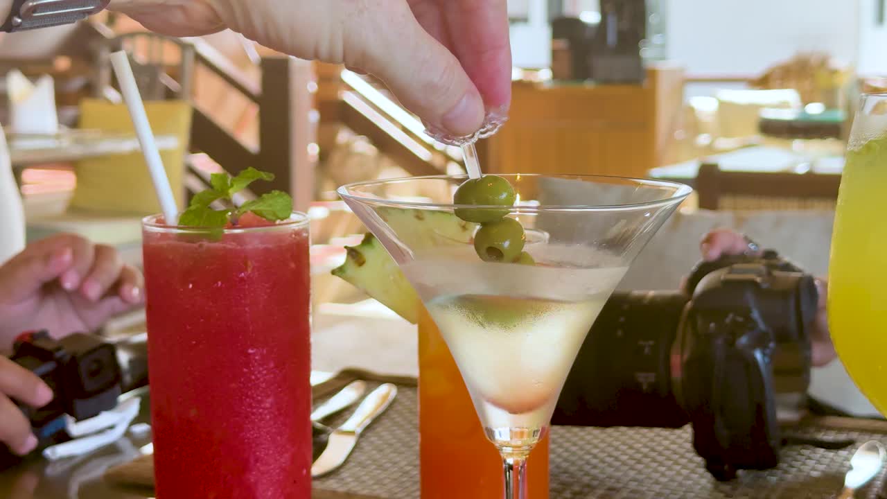 Hand places olive garnish in martini glass beside colorful cocktails, bright daylight, relaxed restaurant setting