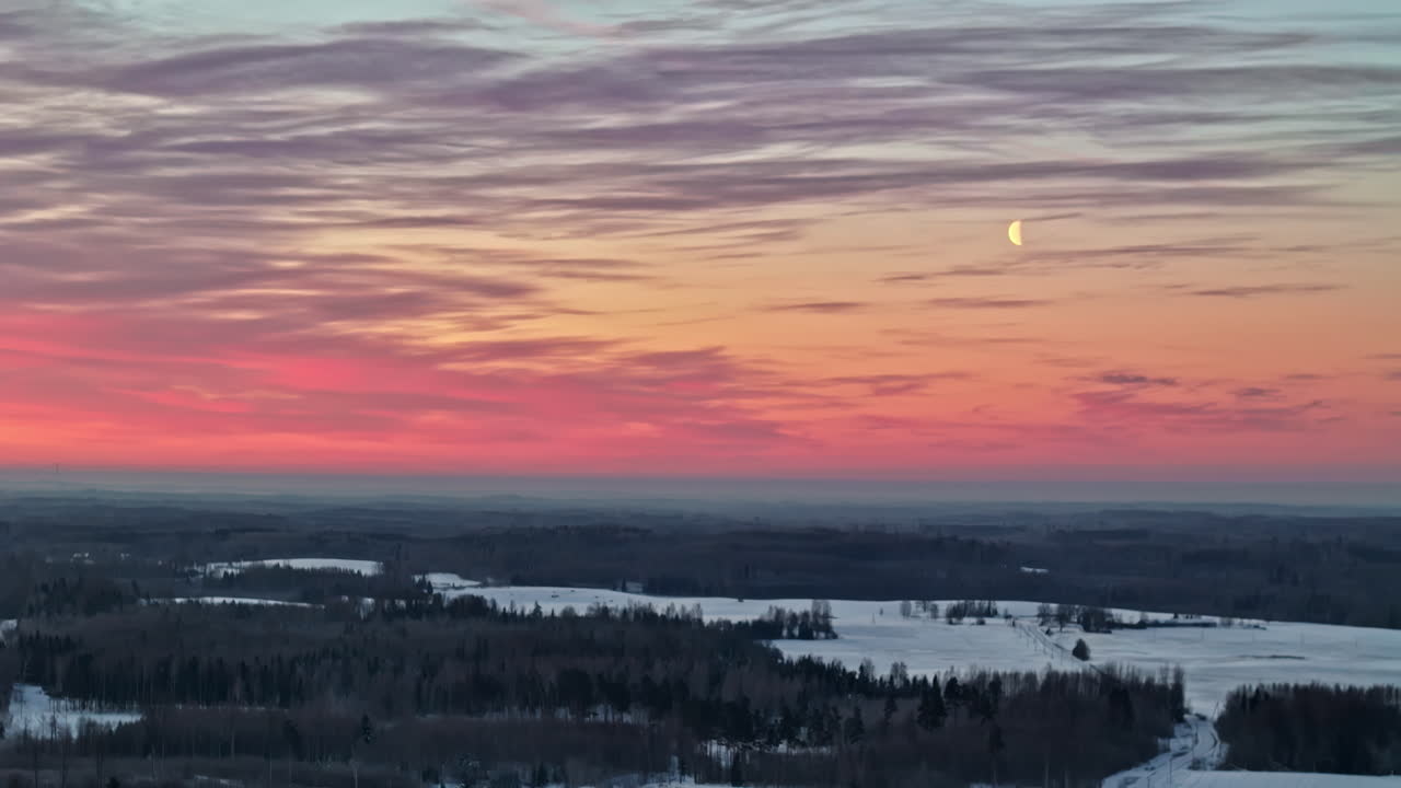 Farverig solnedgangshimmel med en nymåne over en snedækket skov og et frossent landskab