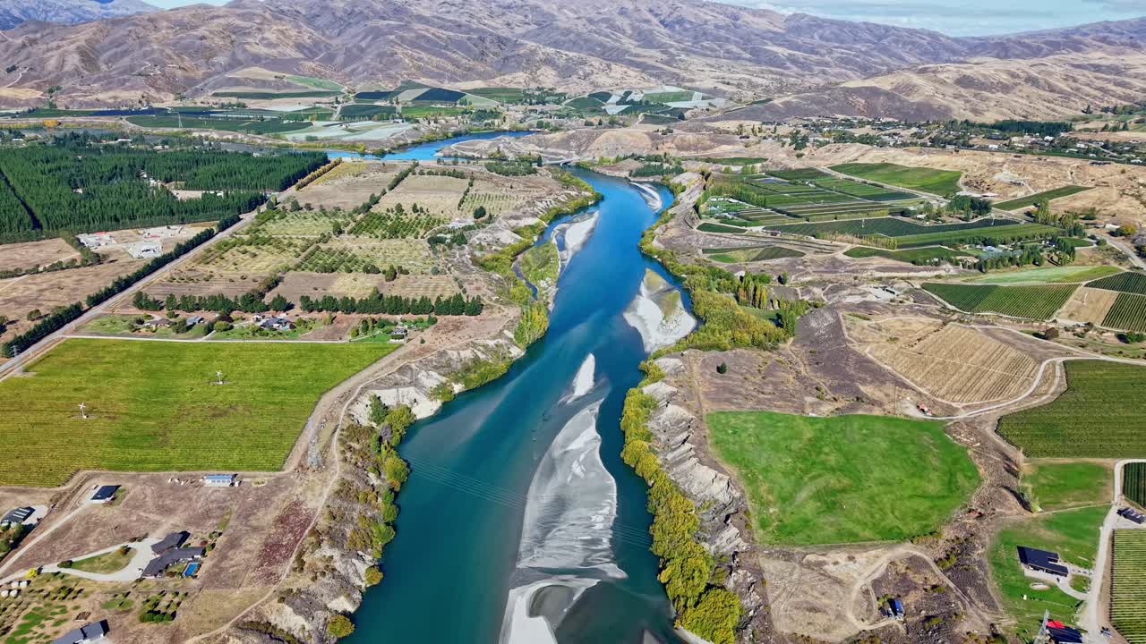 Drone moves backward above the Kawarau River flowing between fields and vineyards near Cromwell, New Zealand