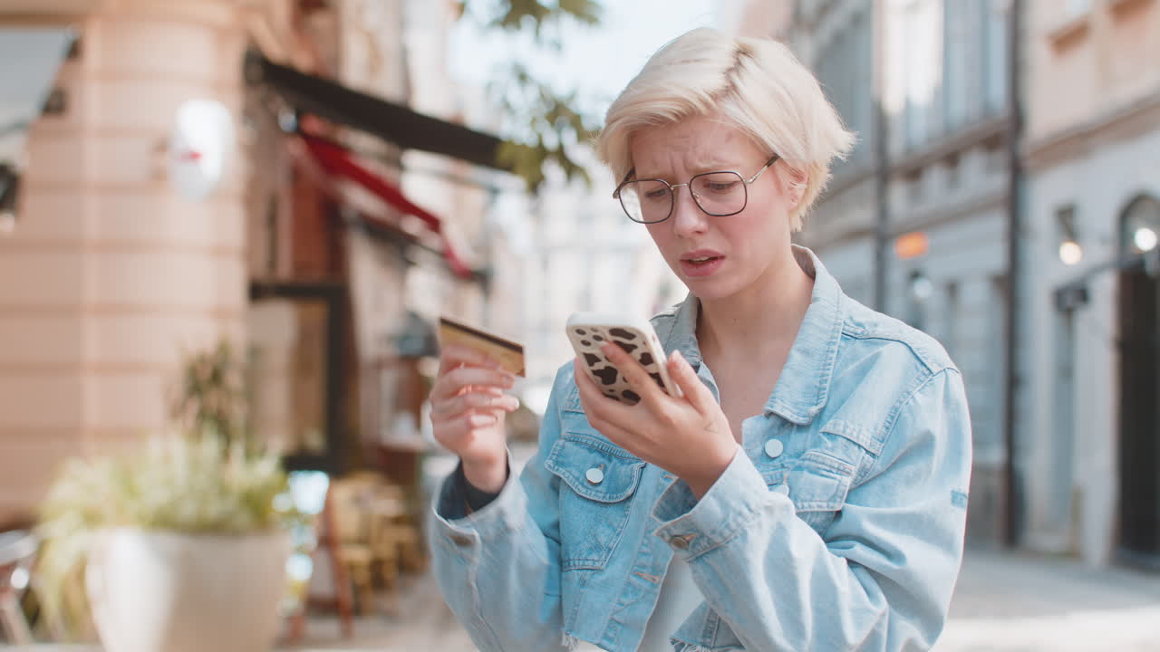 Upset young woman with short blond hair using smartphone and credit card on urban city street