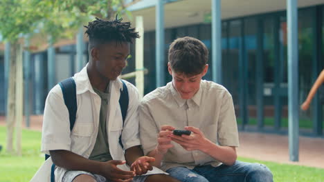 Two-Male-High-School-Or-Secondary-Students-Looking-At-Social-Media-On-Phone-Sitting-Outdoors