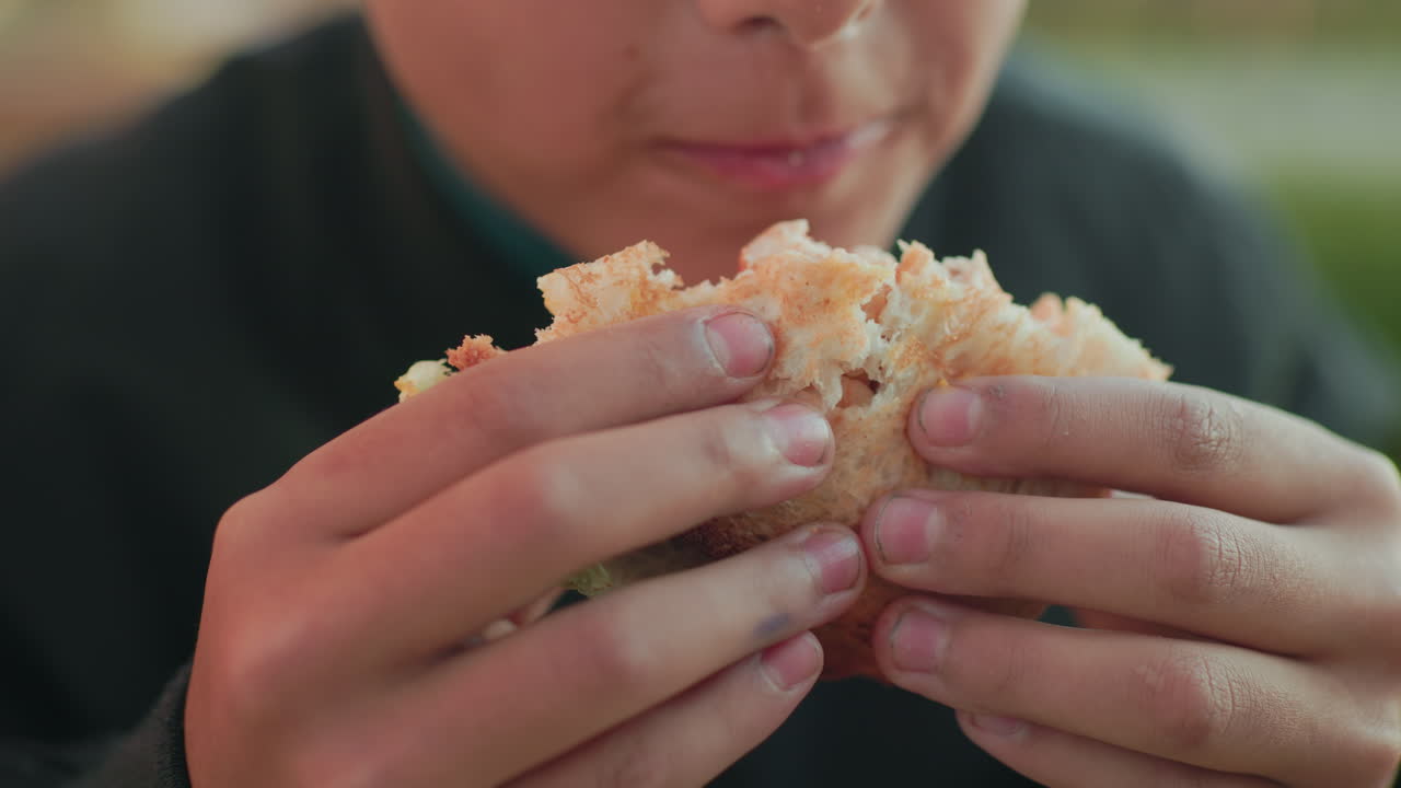 Close up boy savoring burger with both hands, mouth slightly open ready to take bite, showing texture of bread and filling, outdoor daylight scene with blurred background
