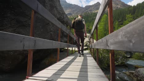 low angle rear view of male hiker crossing wooden bridge in mountain range