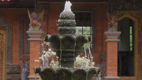 Traditional-stone-fountain-with-flowing-water,-surrounded-by-green-plants,-stands-in-front-of-traditional-Balinese-architecture