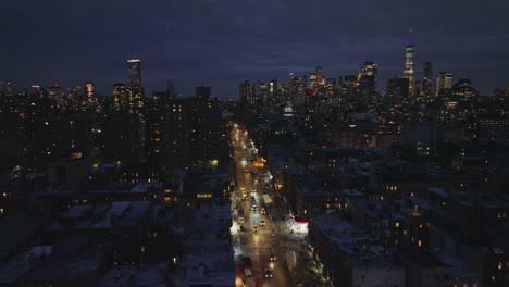 Forwards-fly-above-straight-and-glowing-street-leading-between-apartment-houses-in-urban-neighbourhood.-Night-cityscape-with-downtown-skyscrapers-in-background.-Manhattan,-New-York-City,-USA