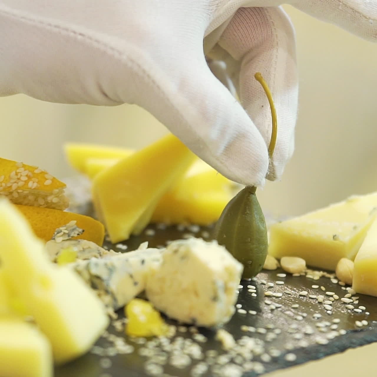A waiter in white gloves holds a black wooden board with pieces of various hard cheese sprinkled with sesame seeds and gently puts green fruit near it. Close-up.