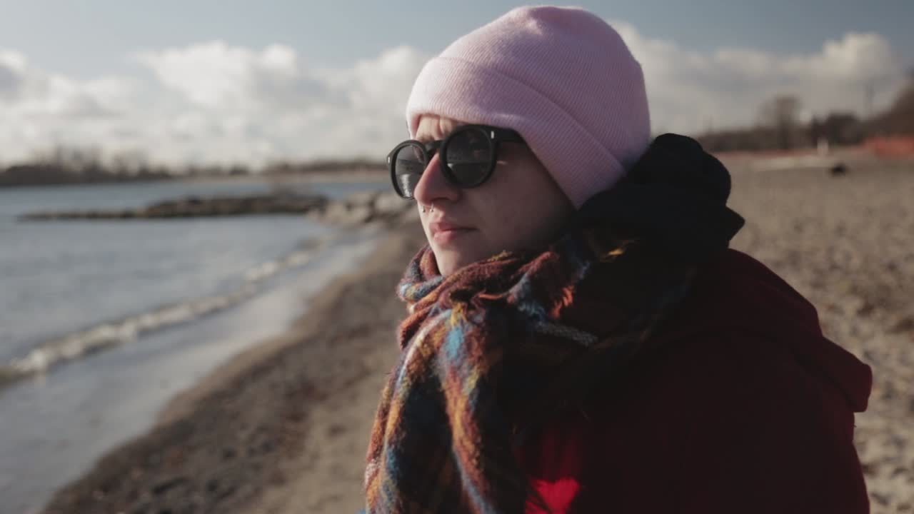 joven turista con chaqueta roja, gafas de sol y gorro rosa parado en la orilla y disfrutando del brillante día de verano - primer plano