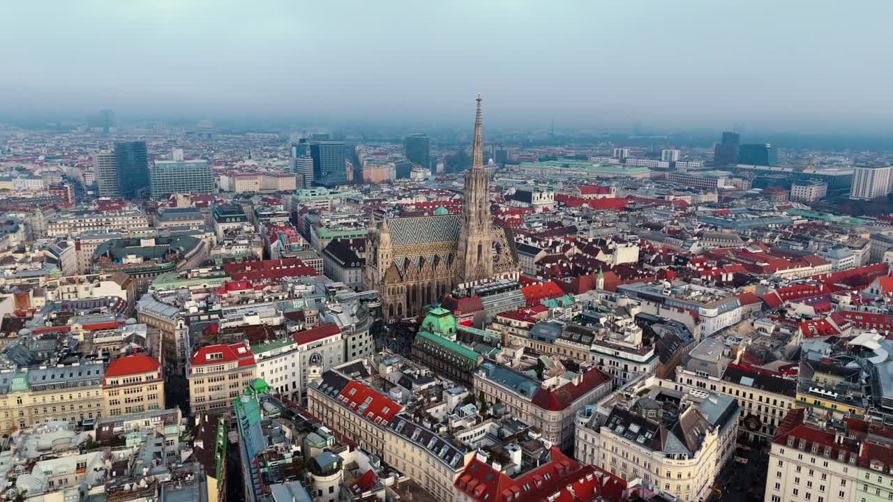 Aerial wide-angle drone shot panning left over St Stephen’s Cathedral in Vienna, showing the patterned roof, surrounding historic rooftops, winter clouds overhead, muted daylight, and tourists visible