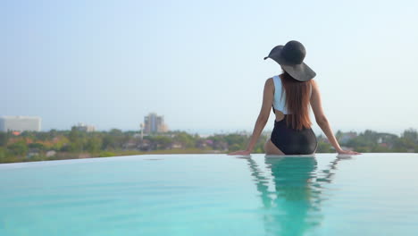 Modelo-Delgado-Con-Traje-De-Baño-Blanco-Y-Negro-Y-Sombrero-Negro-Sentado-Al-Lado-De-La-Piscina-Infinita-De-Espaldas-A-La-Cámara,-Vista-De-La-Ciudad-Al-Fondo