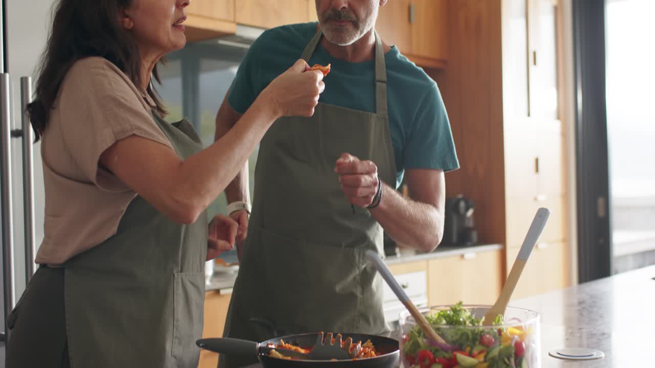 Turning vegetables in pan, couple offering bite and tasting checking flavor at island with aprons