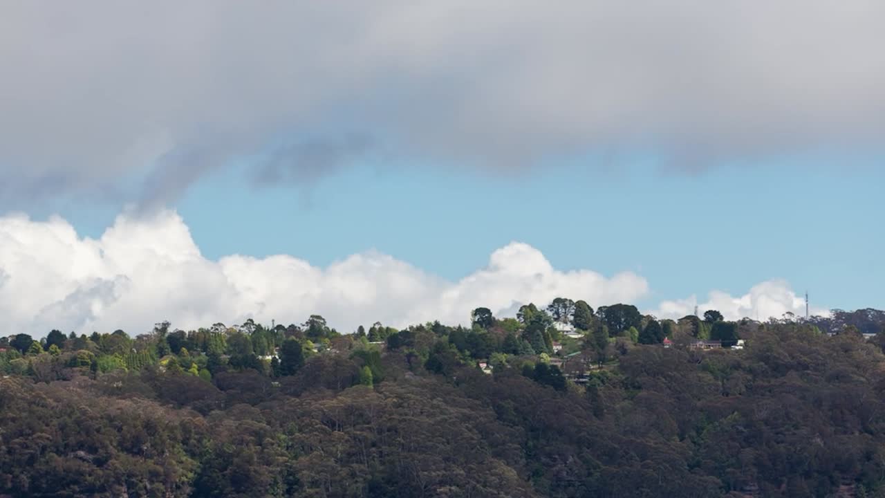 Riprese in time-lapse di un paesaggio scenografico che mostra una collina densamente boscosa in primo piano e un'area residenziale con case sparse sul crinale sullo sfondo. Nuvole bianche fluttuano.