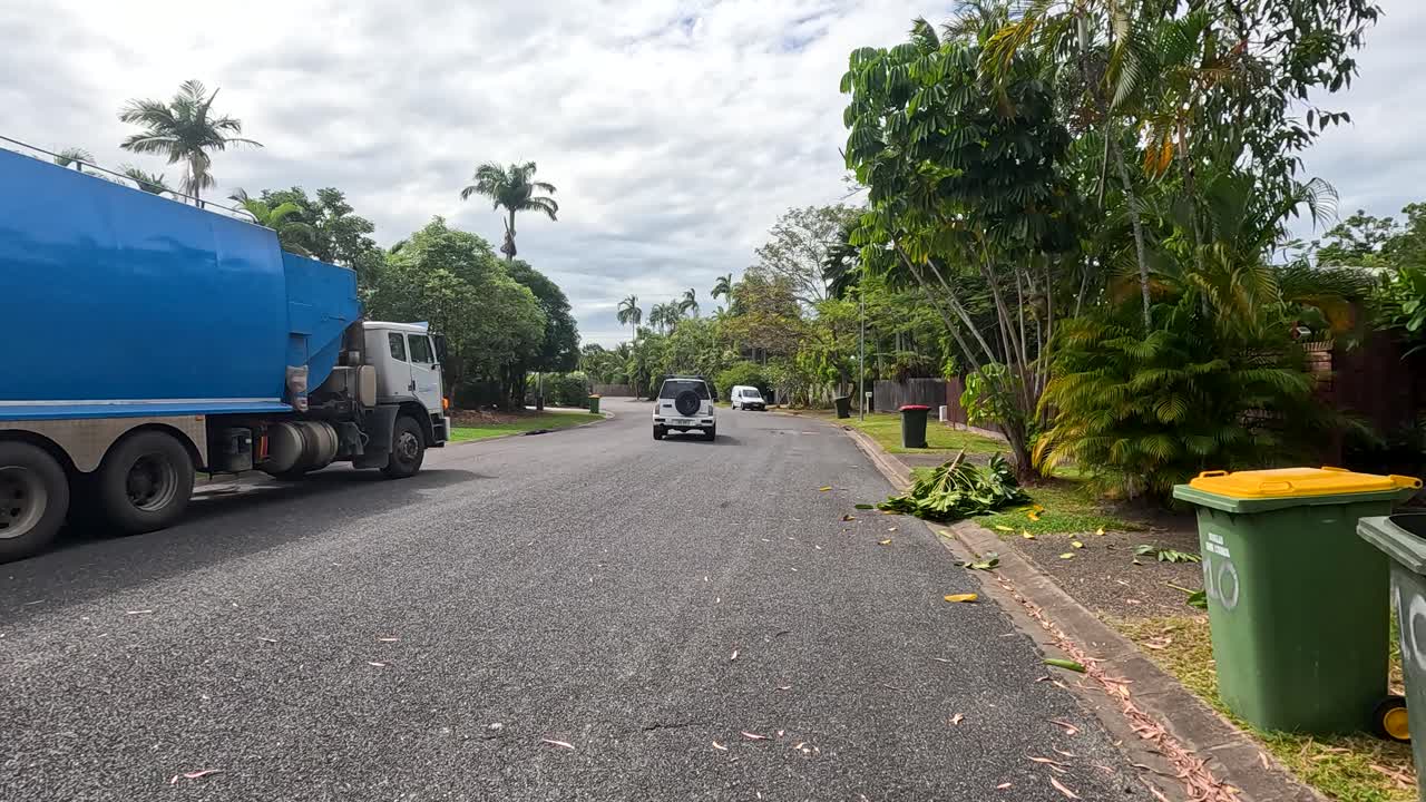 Blue garbage truck and white SUV pass on quiet suburban street with tropical greenery