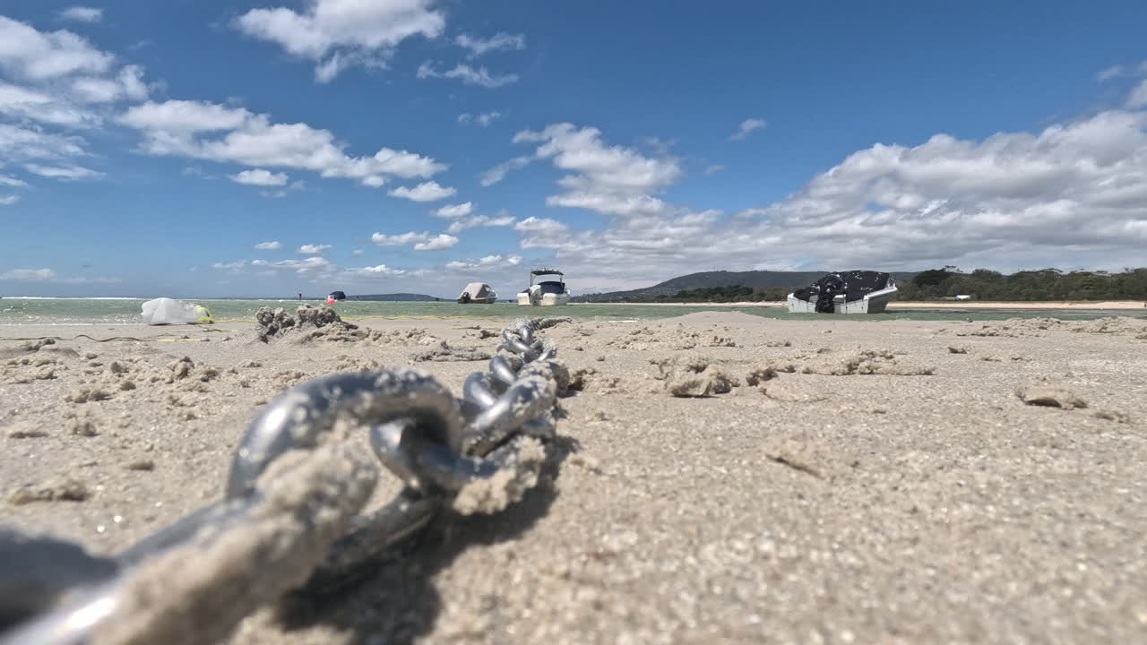 Primo piano di una catena che giace su una spiaggia sabbiosa con acqua, colline distanti e barche galleggianti. Di giorno, con una vista panoramica sullo sfondo.
