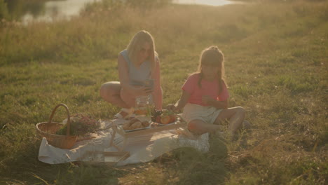 madre e hija sentadas al aire libre compartiendo un picnic relajante bajo la luz del sol, la madre tomando una selfie mientras la hija come frutas frescas. la alfombra de picnic tiene bocadillos, frutas y jarro de agua