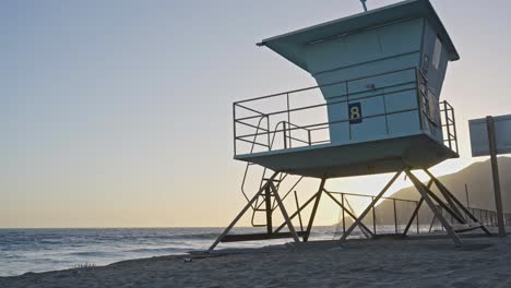 a static shot of a lifeguard tower silhouetted against the setting sun on a quiet california beach, with soft waves rolling in and golden hour light casting long shadows.