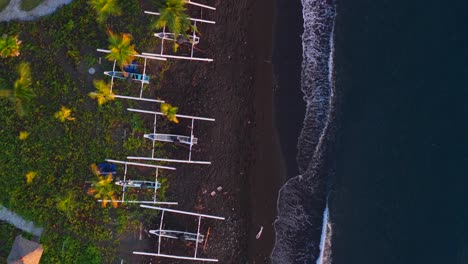 direct overhead aerial of black sand shoreline with lined outrigger boats beside a red roof and foaming waves
