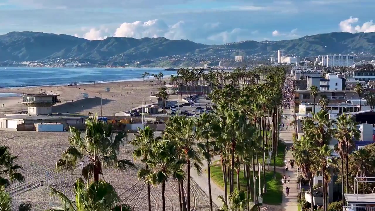 Toma aérea con dron de gran angular giratoria sobre el Ocean Front Walk de Venice Beach, abarcando desde las casas frente a la playa hasta el paseo marítimo bordeado de palmeras con montañas al fondo en un día soleado en Los Ángeles.