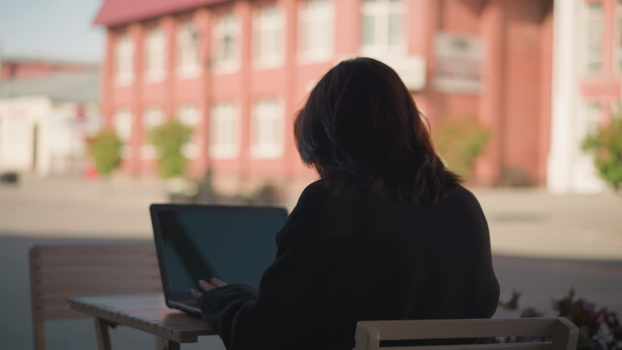 Back view of woman working on laptop outdoors, hair cascading over her shoulders, with a blurred red brick building and greenery in the background