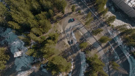 top down aerial following adventurous black suv car driving fast around corner in natural lush pine forest in winter with snow on ground