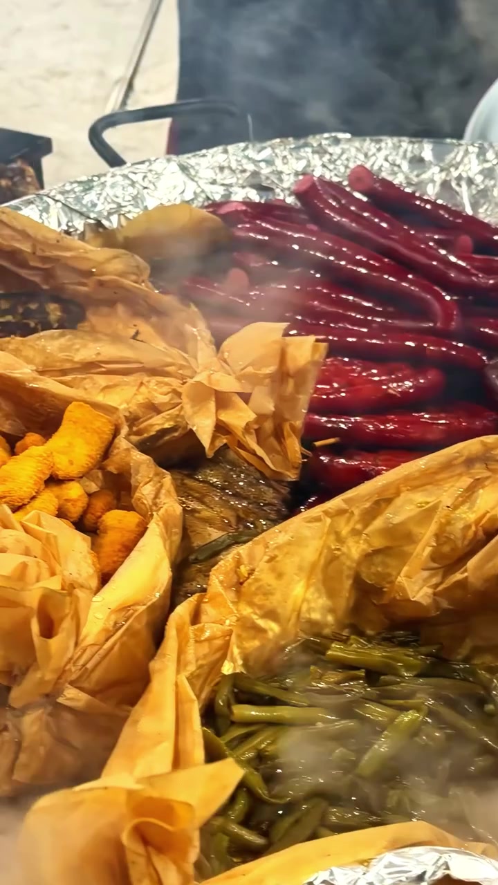Uma variedade de pratos Cajun fumegantes, incluindo frutos do mar fritos, feijão verde e feijão vermelho, cozinhando em travessas de alumínio embrulhadas em papel manteiga ao ar livre.