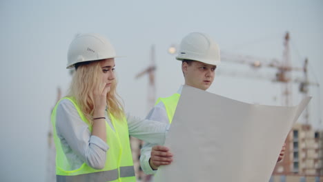 Zwei-Bauarbeiter-Mit-Zeichnungen-Stehen-In-Helmen-Und-Westen-Vor-Dem-Hintergrund-Im-Bau-Befindlicher-Gebäude,-Eine-Frau-Telefoniert-Mit-Einem-Kunden.