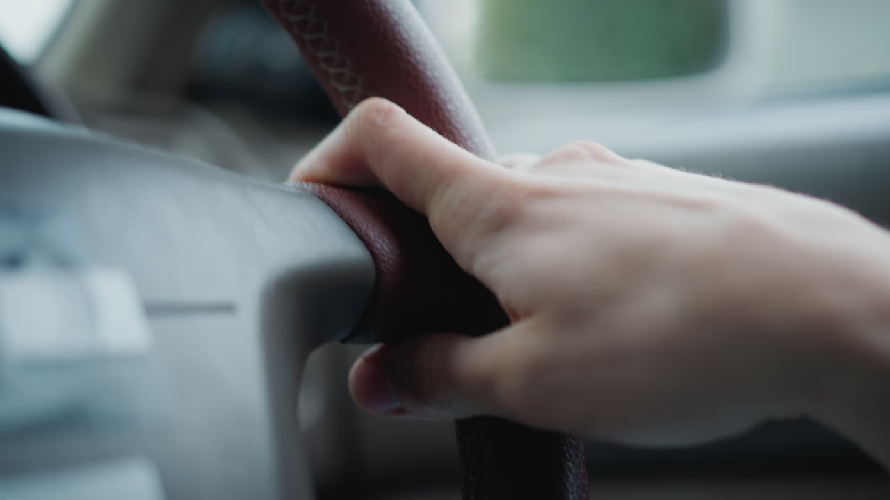 Primer plano de una mano en el volante dentro de un coche, agarre firme en el borde de cuero, salpicadero y ventana borrosos, vegetación exterior que sugiere un trayecto suburbano, conductor conduciendo con concentración, padre chófer