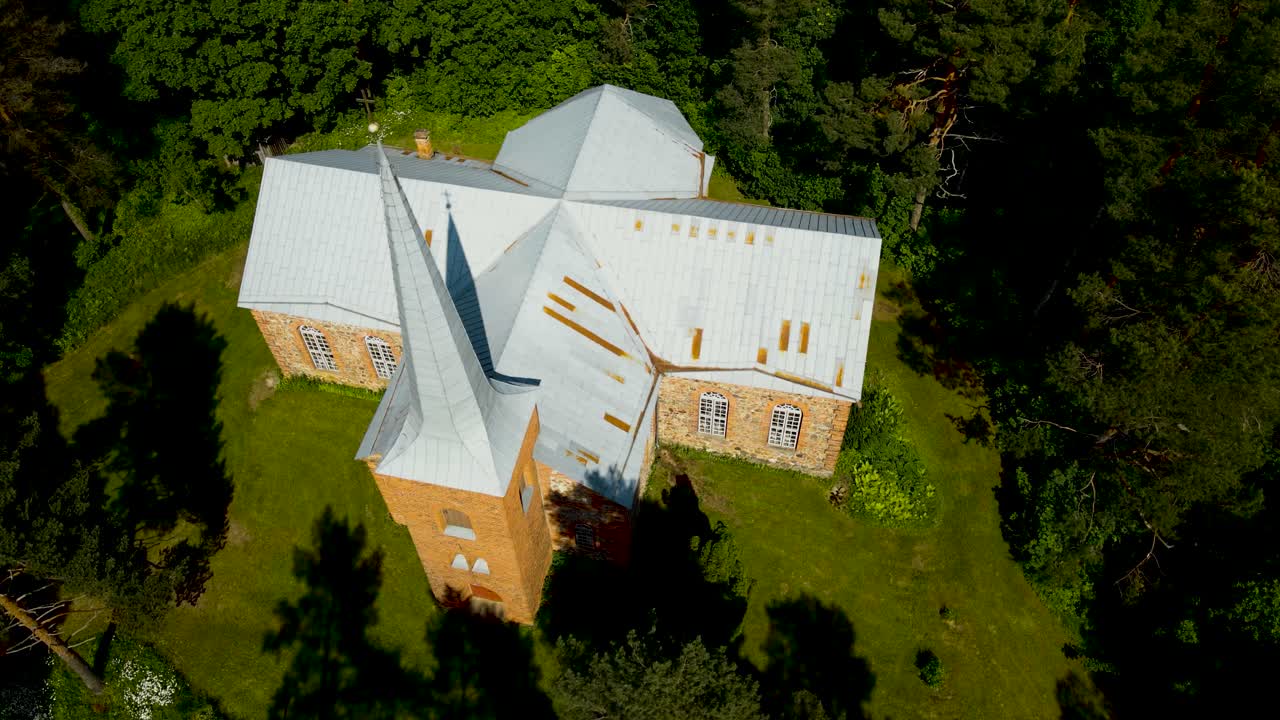 High altitude aerial drone footage orbiting near and over a old red colored brick religious church building during a summer sunny day in Alatskivi Estonia. The church has a tall tower with a cross on