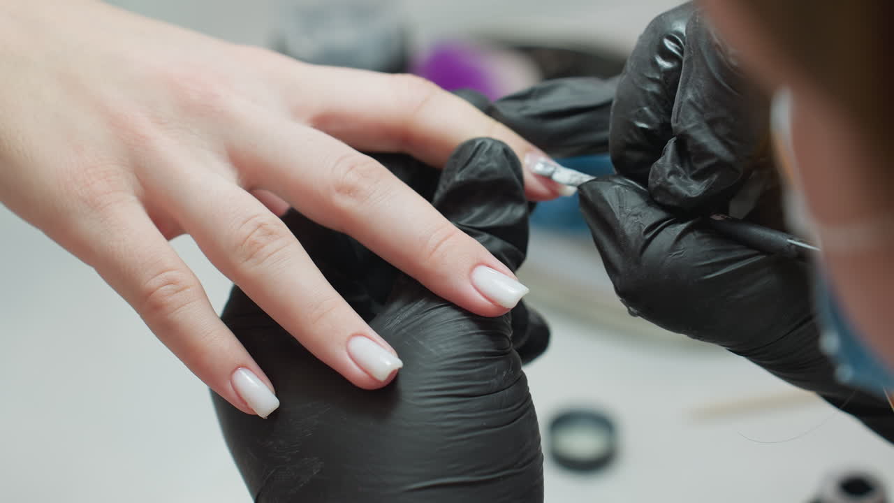 Nail technician wearing black gloves carefully applies white acrylic on client fingernail using brush, focusing on precision and smooth application in clean, professional beauty salon setting