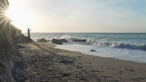hombre juguetón lejos en la playa, arroja piedras, salta, largas sombras se proyectan detrás, las olas del mar se lavan en la costa