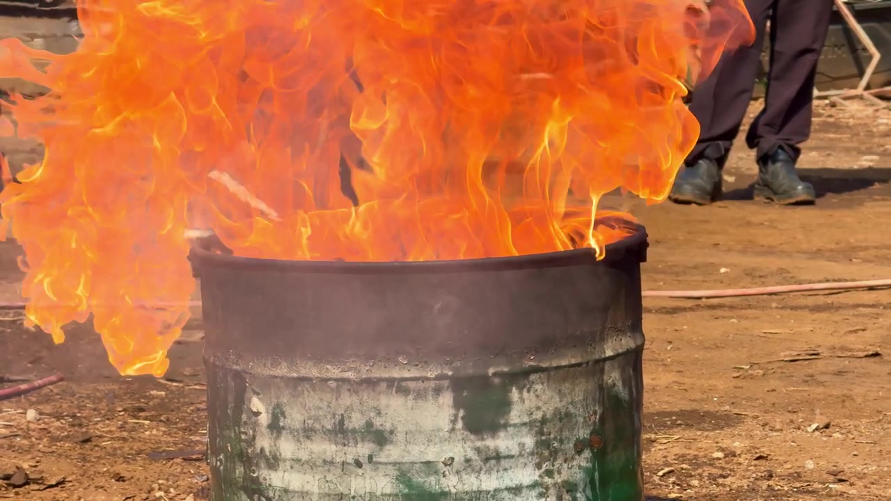 Ildflammer stiger op fra en metaltromle under en udendørs træningsøvelse. Nærbillede af brandforebyggelse, nødøvelse og brandslukningskoncept.