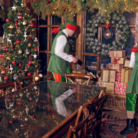 two elves are standing in specially decorated room near the table, holding presents in hands. feast fireplace in the background. christmas spirit concept close up video. christmas eve. new year is coming. elves are putting presents on the table.