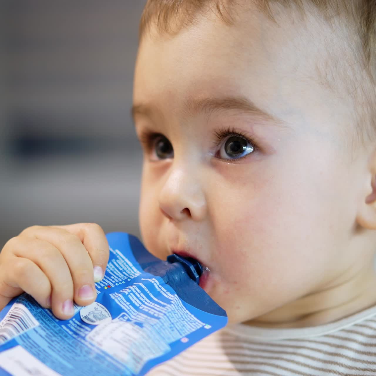 Adorable baby boy holding blue doy pack in mouth. Caucasian toddler eating pureed fruit. Close up. Blurred backdrop