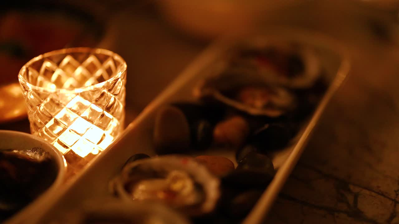 A hand selects oysters by candlelight, creating a warm, intimate dining atmosphere. Soft lighting enhances the luxurious culinary setting