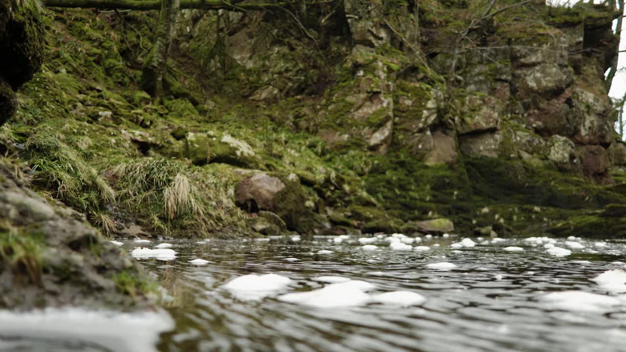 Skumbobler, der flyder på vandets overflade i floden North Esk i Skotland, hvirvler langsomt mod kameraet og ind i et lavt fokusplan med en mosbeklædt klippe i baggrunden.