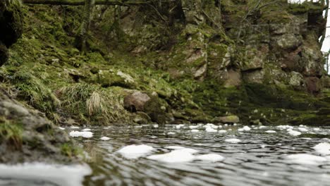 bubbles of foam floating on the surface of the water in the north esk river in scotland slowly swirl towards the camera and into a shallow plane of focus with a moss covered cliff in the background