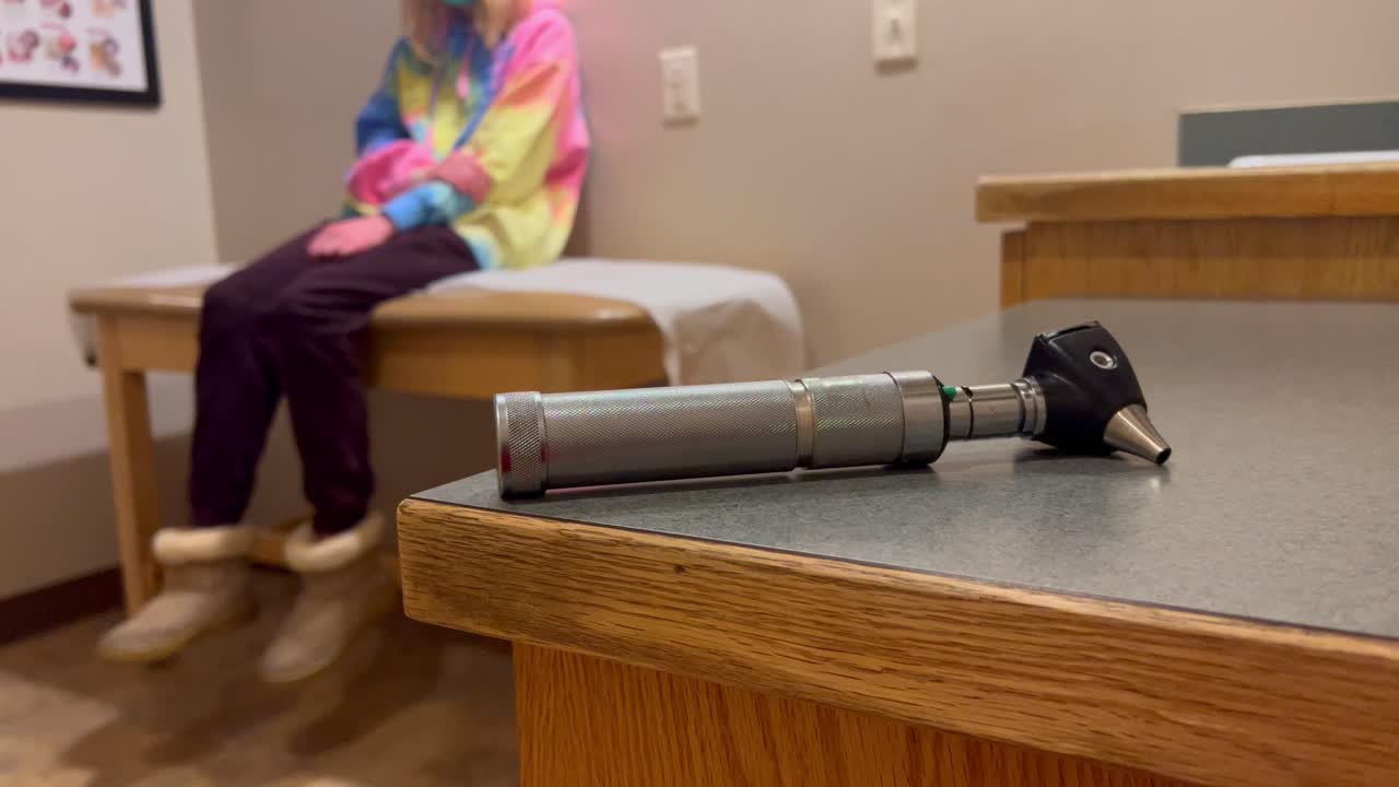 Doctors Otoscope ear nose and throat tool on table with kid child girl or boy in covid-19 mask at Doctor’s office hospital visit waiting on examination exam table nervous fidgeting kicking feet.