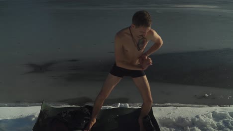 man in shorts standing on black yoga mat on snow covered shoreline of frozen mountain lake exercising and preparing to swim in ice cold water