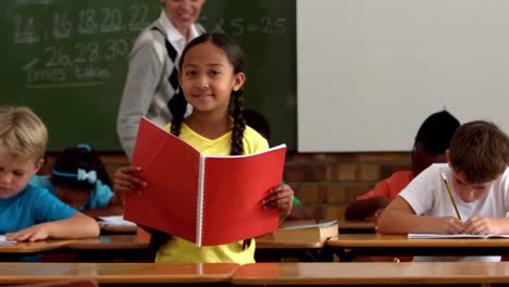 Niña-Sosteniendo-Un-Bloc-De-Notas-Rojo-Sonriendo-A-La-Cámara-En-El-Aula