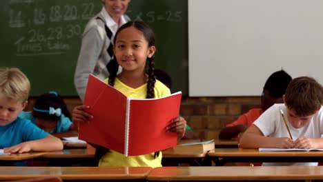 niña con un bloc de notas rojo sonriendo a la cámara en el aula