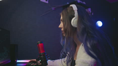 side view of focused young lady with long hair wearing headphones using mouse and keyboard while playing video game at desk with glowing microphone and ambient studio lighting setup