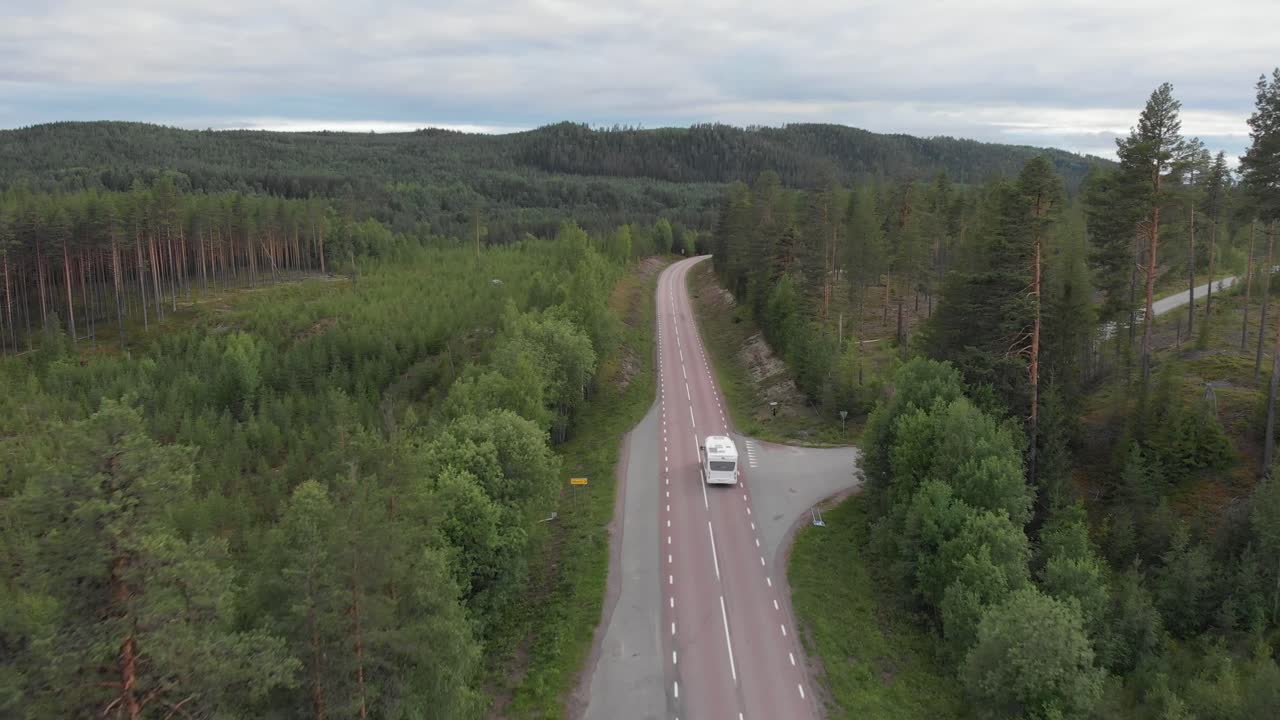 Aerial tracking shot of mountainous lonely road splitting green nordic forest, while chasing white camper van going on an adventure