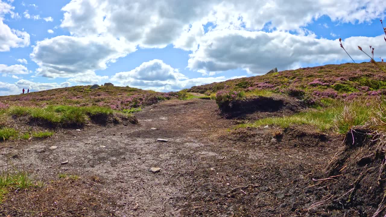 Camera moves steadily up a rugged dirt path bordered by grass and heather under bright daylight, capturing a scenic Scottish hillside with dramatic clouds