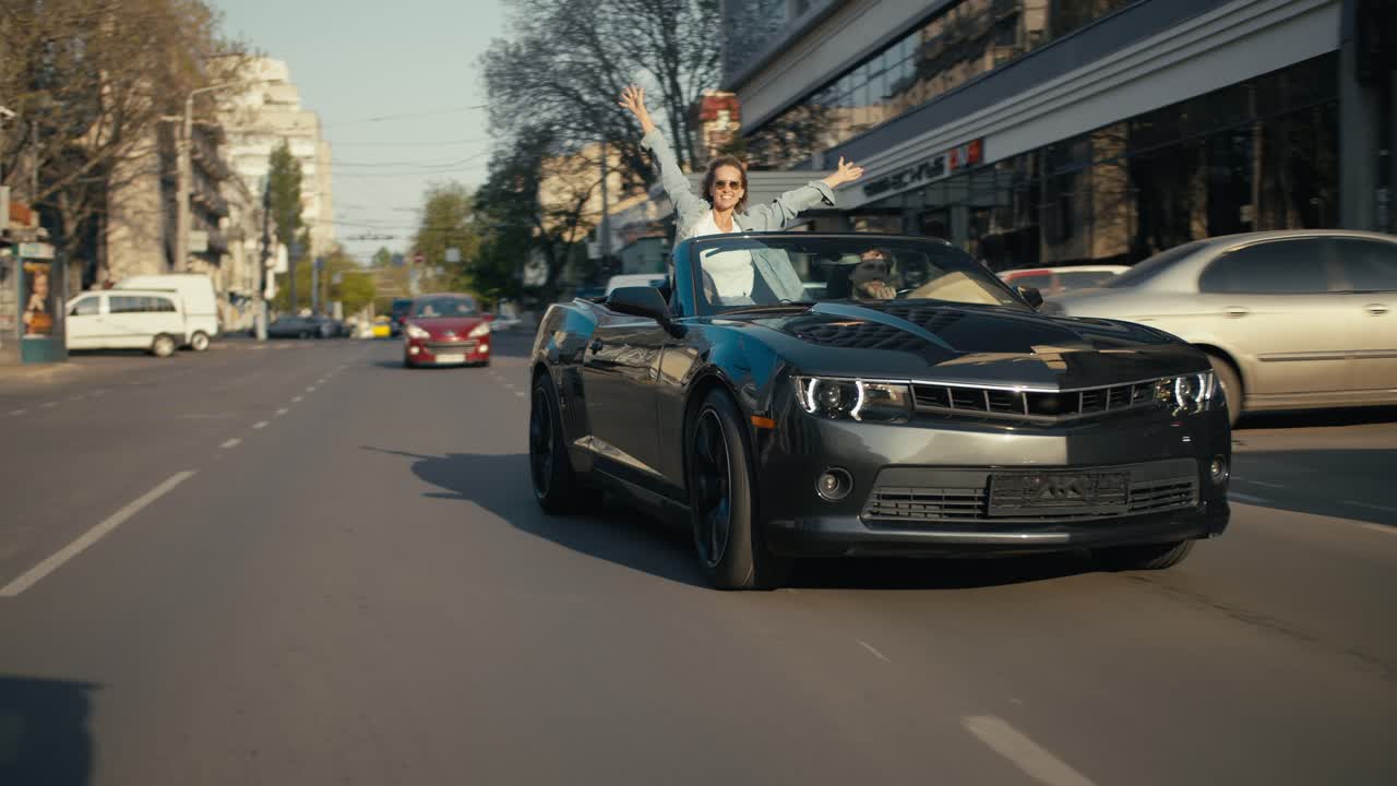 un convertible gris brillante está conduciendo por la calle, una chica rubia en el asiento trasero mira fuera del convertible y extiende los brazos hacia arriba