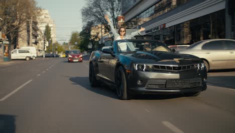 Ein-Glänzend-Graues-Cabrio-Fährt-Die-Straße-Entlang,-Ein-Blondes-Mädchen-Auf-Der-Rückbank-Schaut-Aus-Dem-Cabrio-Und-Streckt-Die-Arme-Nach-Oben