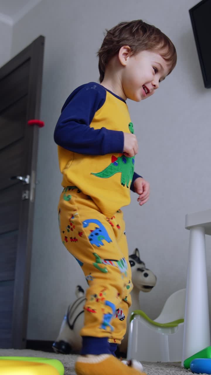 Happy active kid in yellow pajama dances in his room. Baby boy turns around and then falls on his knees. Low angle view. Vertical video